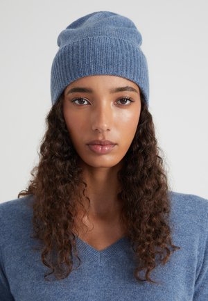Giovane donna con capelli ricci e scuri che indossa un berretto di lana blu e un maglione blu con scollatura a V, che guarda direttamente davanti a sé su uno sfondo neutro.