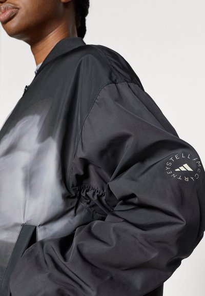 Μαύρο μπουφάν bomber με γυαλιστερό υλικό, φερμουάρ κλεισίματος, ελαστικές μανσέτες και τυπωμένο λογότυπο στο μανίκι. Ομαλή υφή με εφαρμοστή γραμμή.