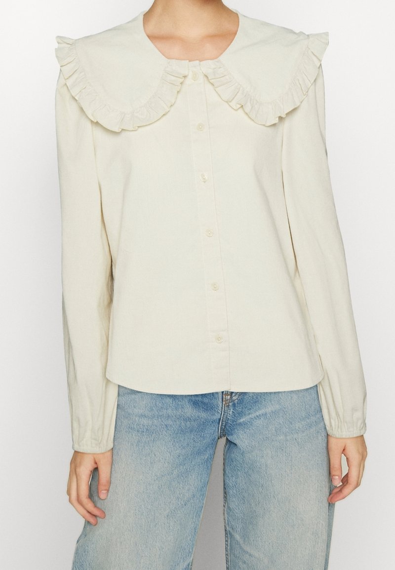 Blouse crème à manches longues avec un col surdimensionné à volants et des boutons sur le devant, portée avec un jean bleu clair par une personne debout.