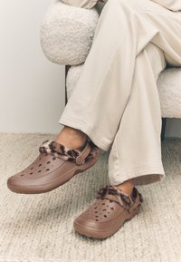 Brown plastic clogs with leopard print faux fur trim and ventilation holes. Worn with light-colored, loose-fitting pants on a textured rug.