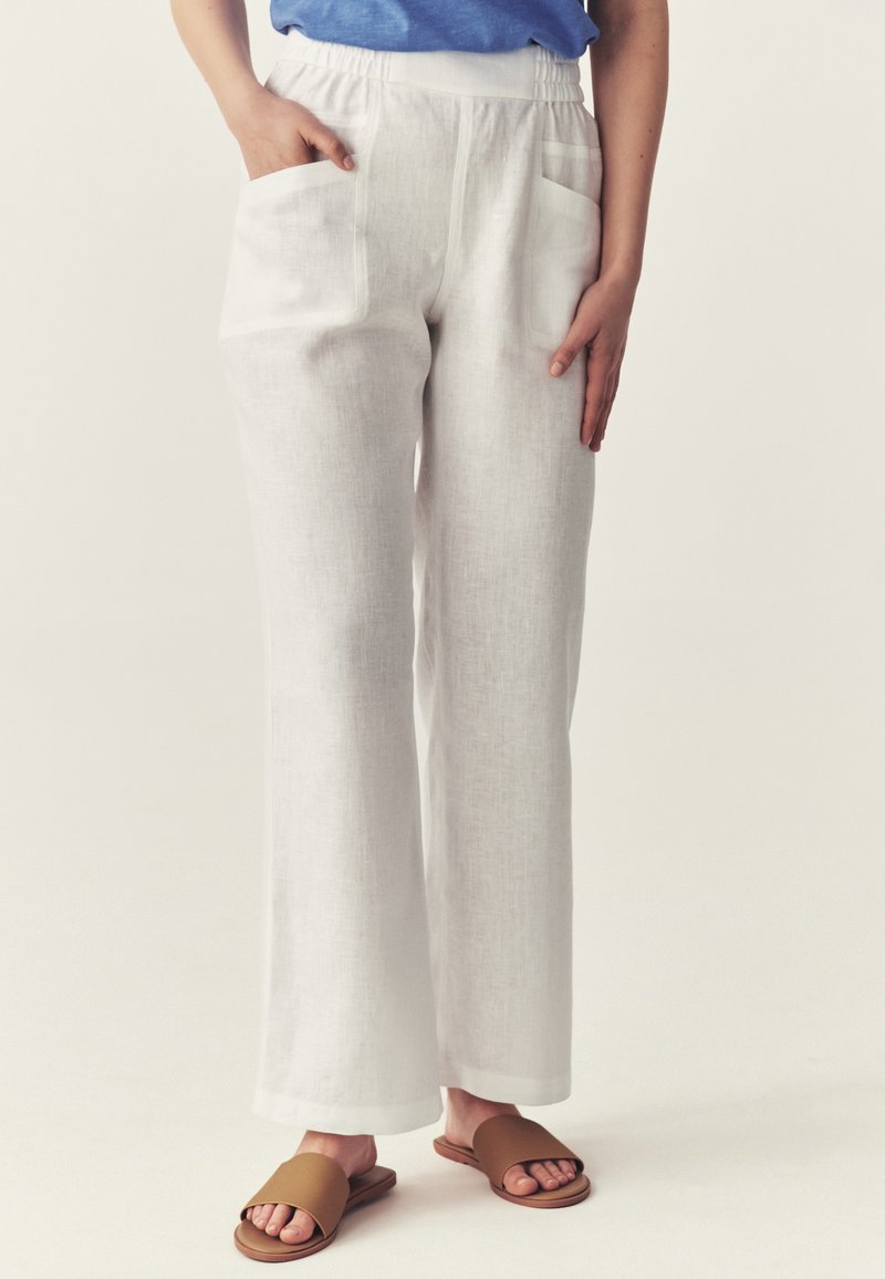 Femme portant un pantalon en lin blanc à jambes larges avec poches avant, un haut bleu et des sandales marron à enfiler, debout sur un fond clair.