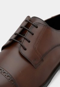 Barna bőr cipő, sima felülettel, fűzős kialakítással és perforált részletekkel az orrán. Lekerekített formájú és kontrasztos varrással díszített.