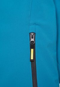 Teal pehmeäkuorinen takkimateriaali, jossa on vetoketjullinen tasku, jossa on hopeanvärinen vetoketju ja musta reunus. Keltainen korostustunnus näkyvissä vetoketjun lähellä.