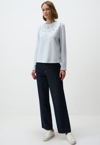 Hellblaues Langarm-Sweatshirt mit dekorativen Verzierungen auf der Vorderseite, kombiniert mit dunkelblauen weit geschnittenen Jeans und weißen Sneakers.