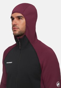 Veste à capuche bordeaux et noire avec une finition texturée, fermeture éclair intégrale et un logo sur la poitrine gauche. Présente un ajustement près du visage.