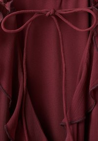 Nahaufnahme einer burgunderfarbenen Bluse aus Stoff mit gebundener Schleife am Ausschnitt und zarten, gerafften Kanten.