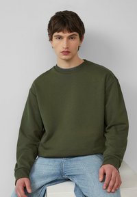 Oliwkowy sweter z okrągłym dekoltem i długimi r�ękawami, o gładkiej fakturze i ściągaczach przy mankietach, w zestawie z jasnoniebieskimi dżinsami.