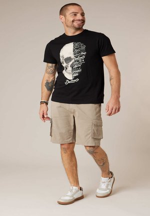 Man met een zwart T-shirt met een schedelprint, beige cargoshorts, witte sneakers en armbanden, staand en glimlachend tegen een effen achtergrond.