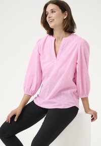Blouse rose clair à enfiler avec un décolleté en V, des manches ballons 3/4 et un motif texturé. Associée à un pantalon noir ajusté.