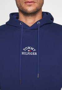 Muž v modré mikině s kapucí s nápisem "Tommy Hilfiger" a malým vyšitým logem červené, bílé a modré vlajky na hrudi.