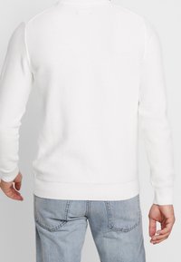 Biały sweter z fakturą i wzorem dzianinowym, okrągłym dekoltem i dopasowanymi mankietami, zestawiony z jasnoniebieskimi dżinsami.