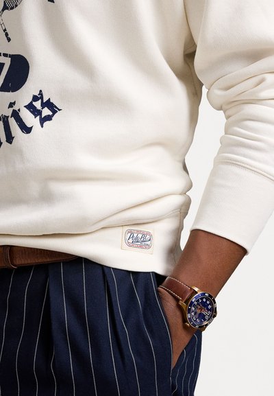 Creme bomulds sweatshirt med marineblå tryk, med et mærke ved kanten. Kombineret med marineblå stribede bukser og et ur med brun læderrem.