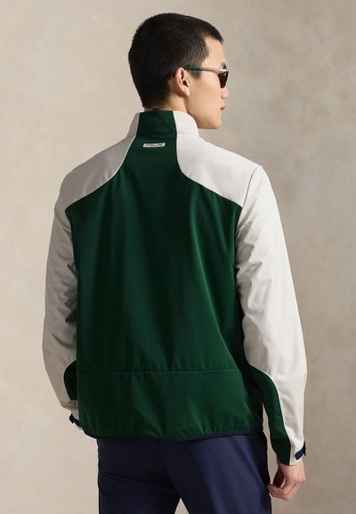 Homme portant une veste bicolore verte et blanche avec un col montant, des lunettes de soleil foncées, et un pantalon bleu marine, debout devant un fond beige uni.