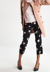 Beżowy trencz na jasnej bluzce, w połączeniu z czarnymi krótkimi spodniami z białymi kwiatowymi wzorami. Czarne szpilki dopełniają całość.