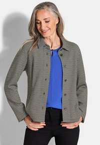 Grijze jas met een houndstooth-patroon en donkere knopen, met een kraag en een knoopsluiting, gedragen over een blauwe top en zwarte jeans.