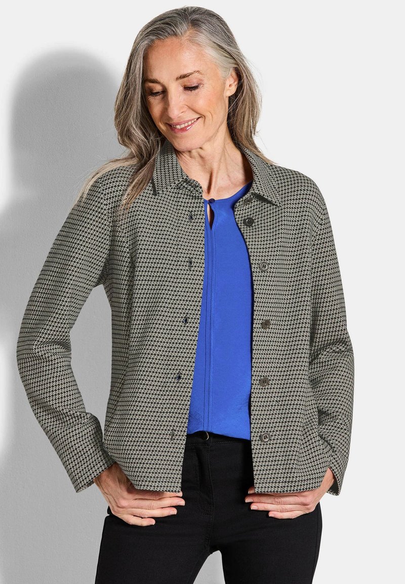 Grijze jas met een houndstooth-patroon en donkere knopen, met een kraag en een knoopsluiting, gedragen over een blauwe top en zwarte jeans.