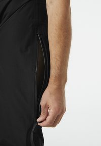 Zwarte broek met een zijrits, gladde stof en een slim fit snit, met een zichtbare hand die de ritstrekkering vasthoudt.