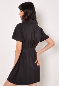 Robe noire avec des manches courtes, un col, et une taille ceinturée. Caractérisée par un dos froncé pour ajouter du volume et de l'élégance.