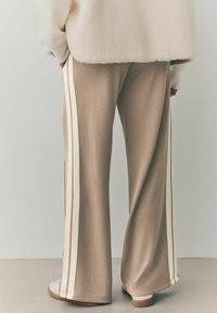 Pantalons larges beiges en tissu doux avec des bandes latérales crème. Présentent une coupe décontractée et des ourlets à revers, associés à des chaussures blanches.