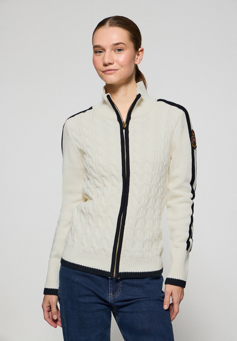 Krämfärgad kabelstickad tröja med hög krage, dragkedja och marinblå detaljer på ärmar och nederkant. Har ett dekorativt märke på ärmen.