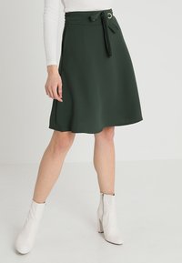 Une jupe verte foncée, arrivant au genou, fabriquée à partir d'un tissu léger, avec une taille à lien, un accent en matériel circulaire et une texture lisse.
