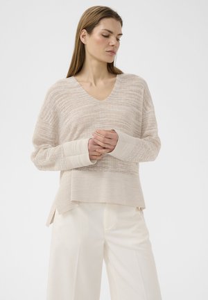 Femme aux longs cheveux portant un pull en maille beige à col en V et un pantalon blanc à jambes larges, debout, les mains jointes, regardant vers le bas.