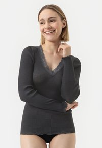 Glückliche vrouw met kort blond haar, gekleed in een donkergrijs ribbed langemouwen shirt met kant aan de V-halslijn, tegen een witte achtergrond.