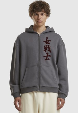 Sweat à capuche zippé gris en coton doux, présentant un grand design graphique texte rouge foncé à l'avant, avec des poches avant et une capuche.