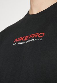Musta Nike Pro -t-paita, jossa on punainen ja valkoinen logoteksti. Valmistettu sileästä kankaasta, jossa on pyöreä pääntie ja lyhyet hihat.