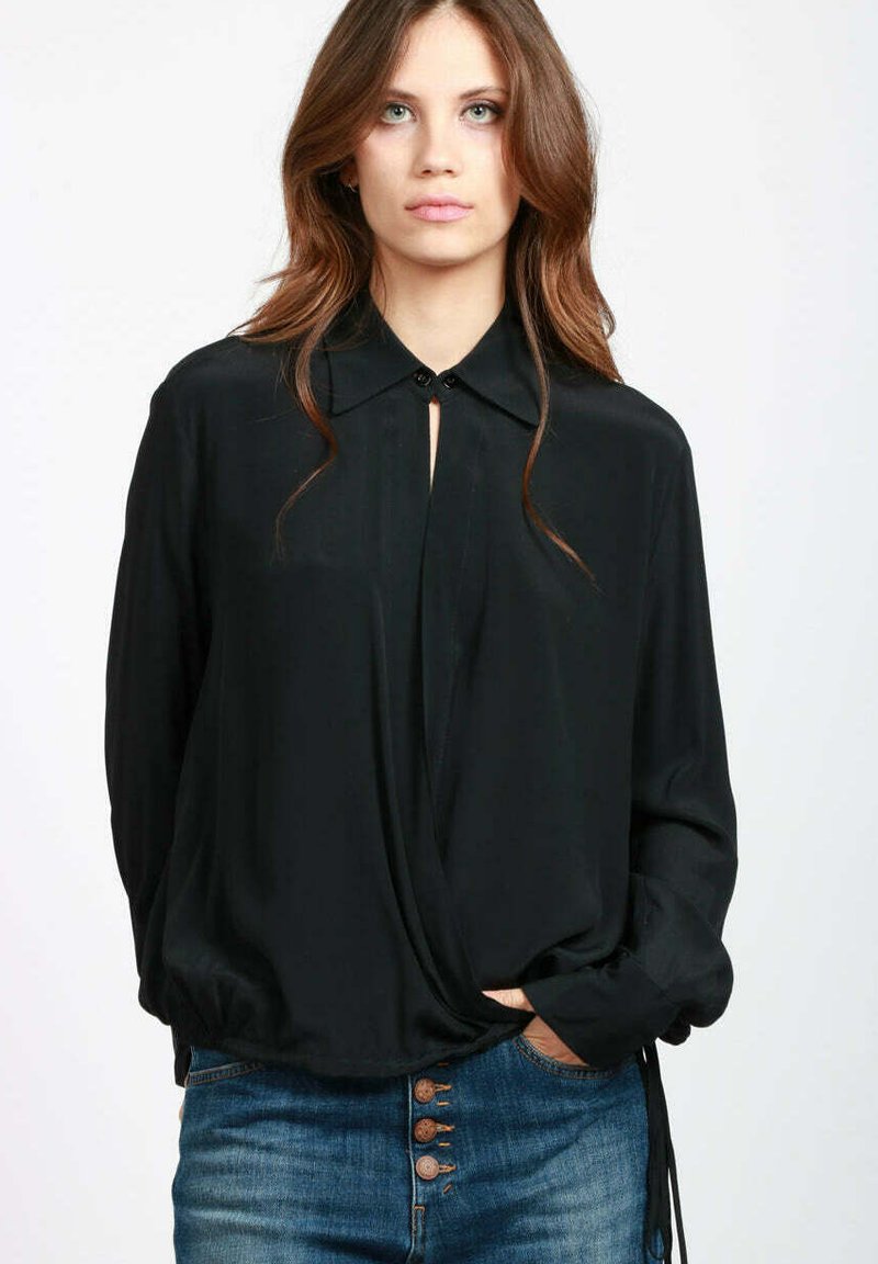 Blusa nera a maniche lunghe con colletto a punta, scollatura a goccia e dettagli con lacci regolabili ai polsini, indossata con jeans blu.
