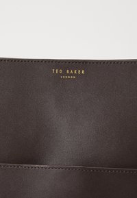 Ruskea keinonahkainen lompakko, jossa on sileä pinta. Varustettu kohokuvioidulla "TED BAKER" -logolla kultaisena. Alhaalla näkyvät kaksi pystysuuntaista korttipaikkaa.