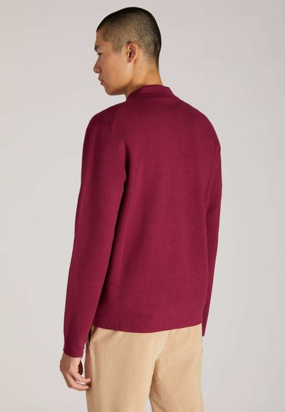 Polo a maniche lunghe bordeaux con finitura testurizzata, dotato di colletto classico e spacchi laterali, abbinato a pantaloni in corduroy di colore chiaro.