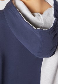Personne portant un sweat à capuche bleu marine superposé à un t-shirt gris clair, montrant la capuche repliée et le haut du dos.