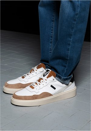 Voeten met witte en bruine veter sneakers met een spijkerbroek op een lichtgrijze betegelde vloer.