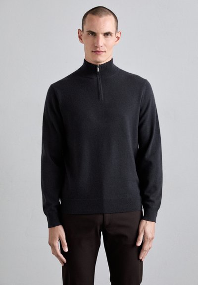 Pull noir à col zippé en tissu doux. Dispose de manches longues, de poignets et d'un ourlet côtelés, ainsi que d'une texture lisse. Porté avec un pantalon marron.