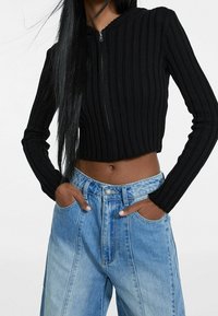 Svart ribbad croppad jacka med dragkedja och långa ärmar, bärs med högmidjade urtvättade blå jeans. Händerna är placerade i fickorna.