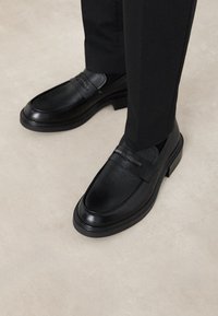 Mocassins en cuir noir au fini lisse, avec une sangle décorative et un petit talon épais. Portés avec un pantalon noir ajusté.