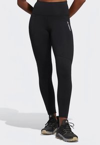 Nohy oblečené v čiernych športových legínach s vysokým pásom a čiernych trailových bežeckých topánkach Adidas na svetlom pozadí.