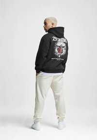 Must ja sweatshirt, mille seljal on graafiline kujundus kotkasta, selga tõmmatud valge särke peale, koos heledate hallide dressipükste ja valgete kingadega.