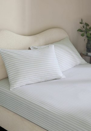 Bett mit cremefarbener, geschwungener Kopfstütze, weißem Spannbettlaken und Kissen mit hellgrauen horizontalen Streifen sowie einem kleinen Glastisch mit einer grünen Pflanze.