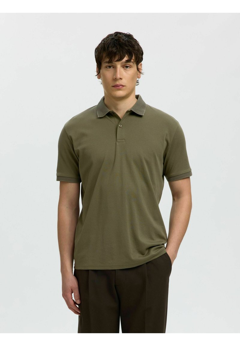 Jeune homme aux cheveux courts et foncés, portant un polo olive à manches courtes et un pantalon foncé, debout devant un fond clair uni.