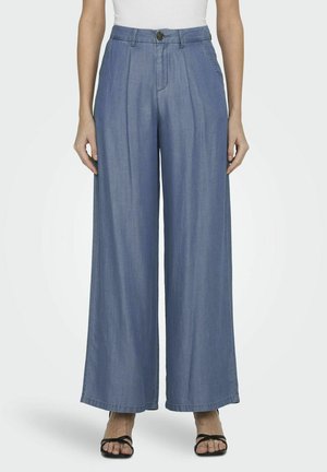 Vrouw in hoge taille, wijde blauwe denim broek met zijzakken, gecombineerd met zwarte gespen sandalen en een witte top.