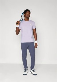 Lichtpaars sportief T-shirt met een klein logo, gecombineerd met donkergrijze sportbroek en witte sneakers. De persoon houdt een tennisracket vast.