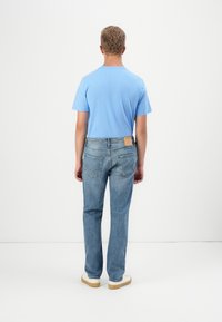 Moški stoji s hrbtom obrnjen, oblečen v svetlo modro majico, modre kavbojke z usnjenim pečatom in bele superge v prazni beli sobi.