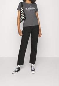 Szara bawełniana koszulka z logo "HOLLISTER", połączona z czarnymi dżinsami i czarnymi wysokimi sneakersami. Towarzyszy jej dekoracyjna torba w stylu siatki.