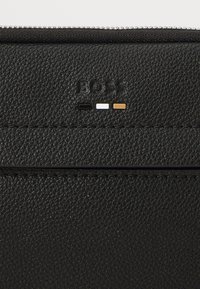 Black textured leather wallet featuring embossed brand logo, a zip closure, and three color accent stripes: black, white, and gold.