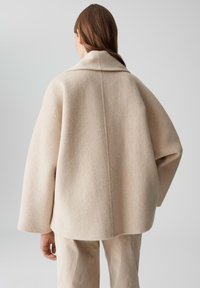 Vrouw met lang bruin haar, gekleed in een oversized beige wollen jas en bijpassende broek, gezien vanuit de rug tegen een effen grijze achtergrond.
