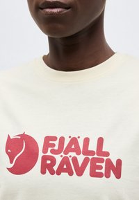 Άτομο που φοράει ανοιχτόχρωμο πουκάμισο με κόκκινο λογότυπο Fjällräven που απεικονίζει μια στυλιζαρισμένη κεφαλή αλεπούς και έντονο κείμενο στο στήθος.