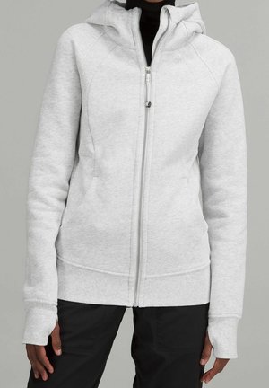 Person iført en lysegrå lynlåshoodie med tommelhuller på ærmerne og sorte bukser, stående mod en ensfarvet grå baggrund.