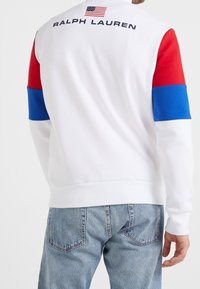 Bílá mikina s červenými a modrými rukávy, s logem a vlajkou na zádech. Měkká látka a žebrované manžety. Ležérní design.